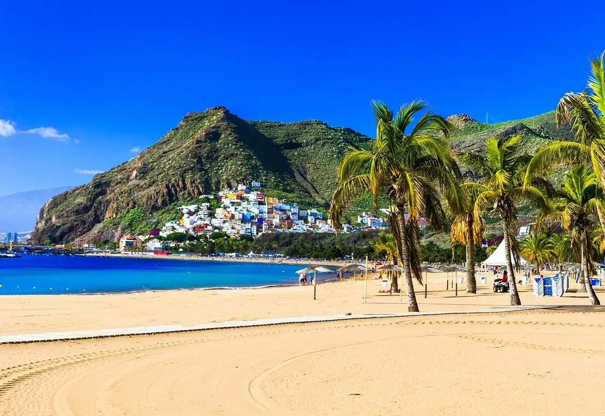 Playa de arena dorada con palmeras y un pueblo colorido en la ladera de una montaña. ¡Un destino ideal donde viajar en agosto barato!