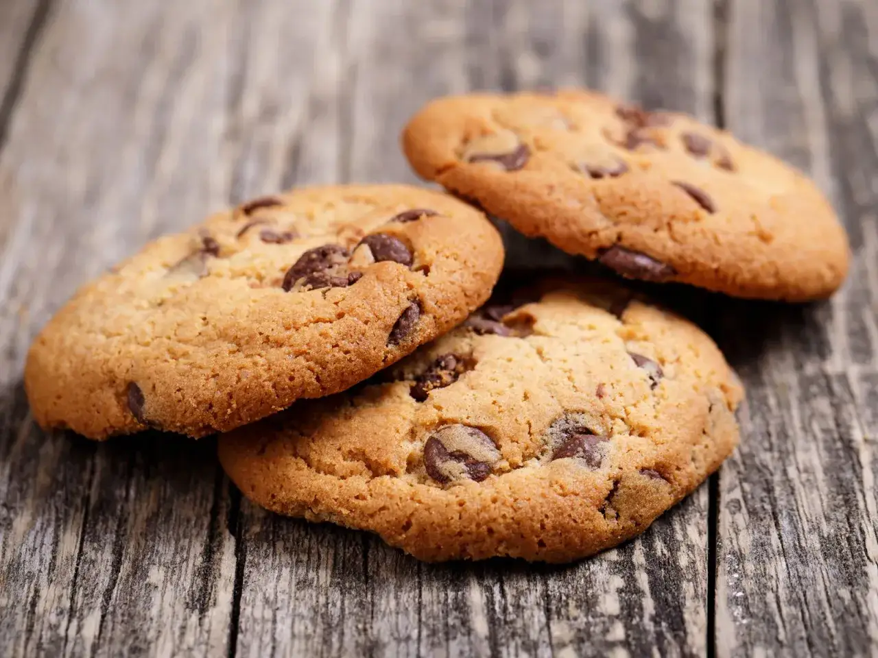 Drei köstliche, goldbraune glutenfreie Kekse mit Schokoladenstückchen liegen auf einer rustikalen Holzoberfläche.
