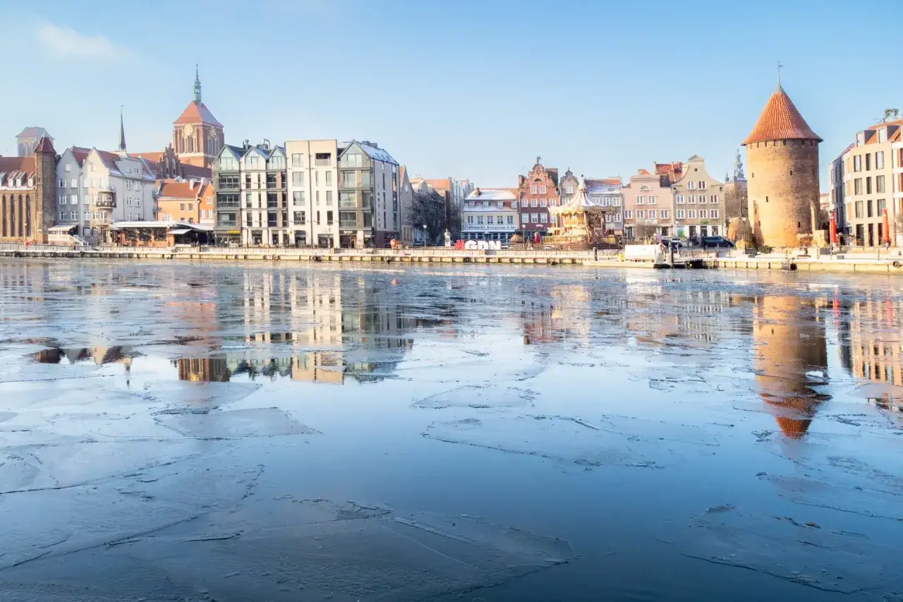 Zamarznięta rzeka w Gdańsku, z widokiem na zabytkową zabudowę Starego Miasta i charakterystyczną basztę.