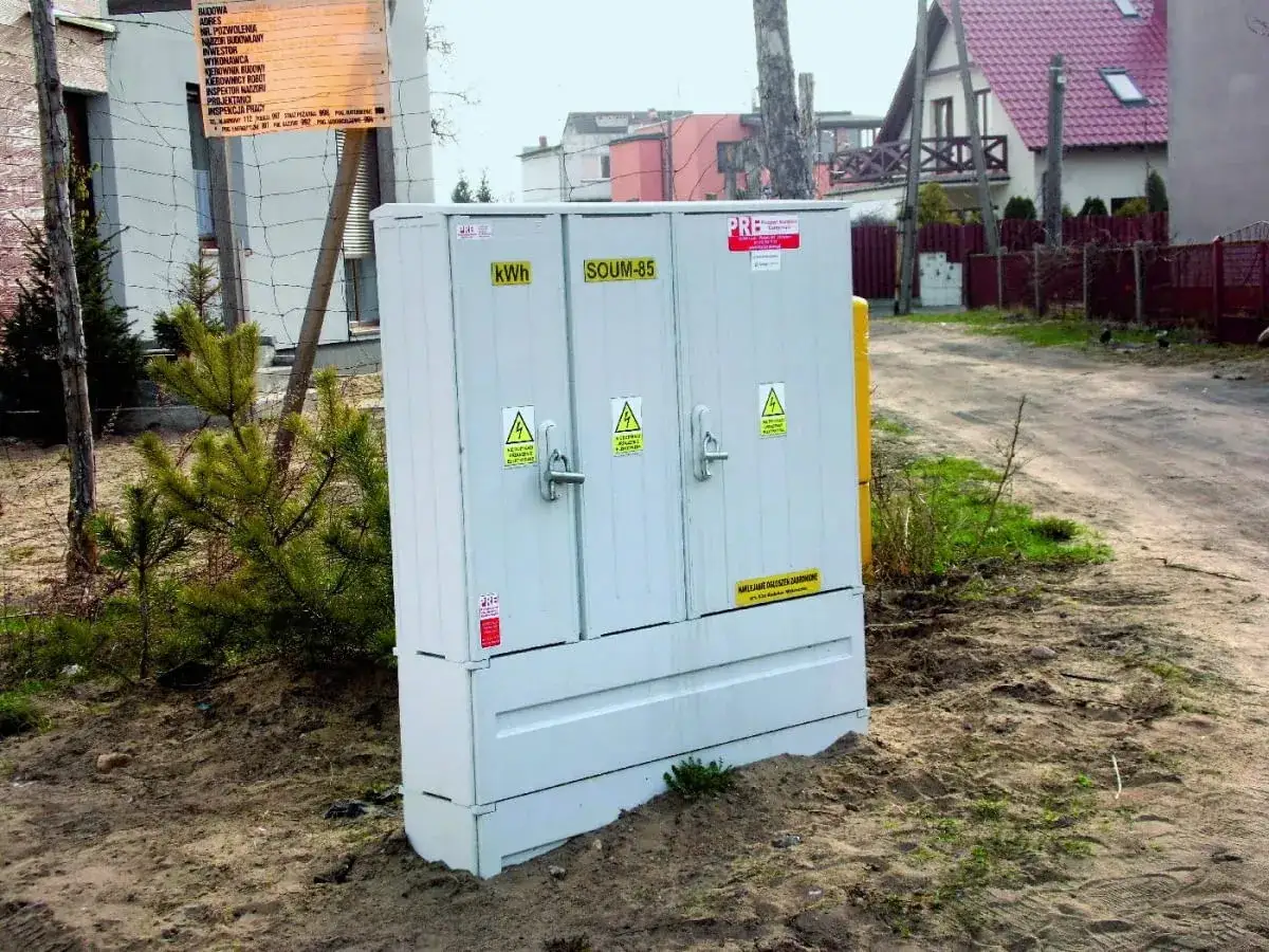 Skrzynka elektryczna Tauron do przyłączenia prądu do działki. W tle domy i droga.