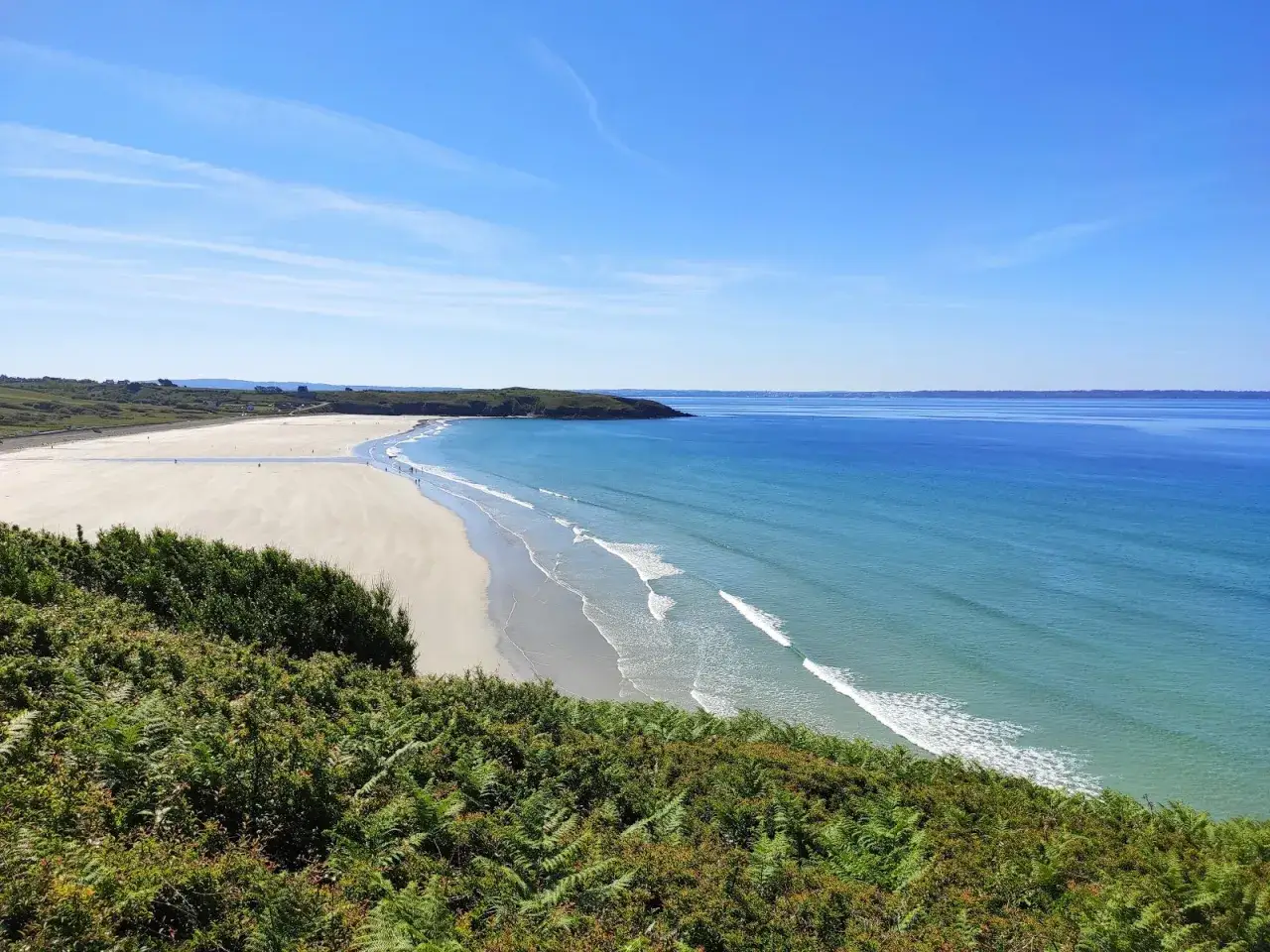 Vue panoramique d'une plage trez bellec, avec sable clair, mer bleue et végétation luxuriante au premier plan.