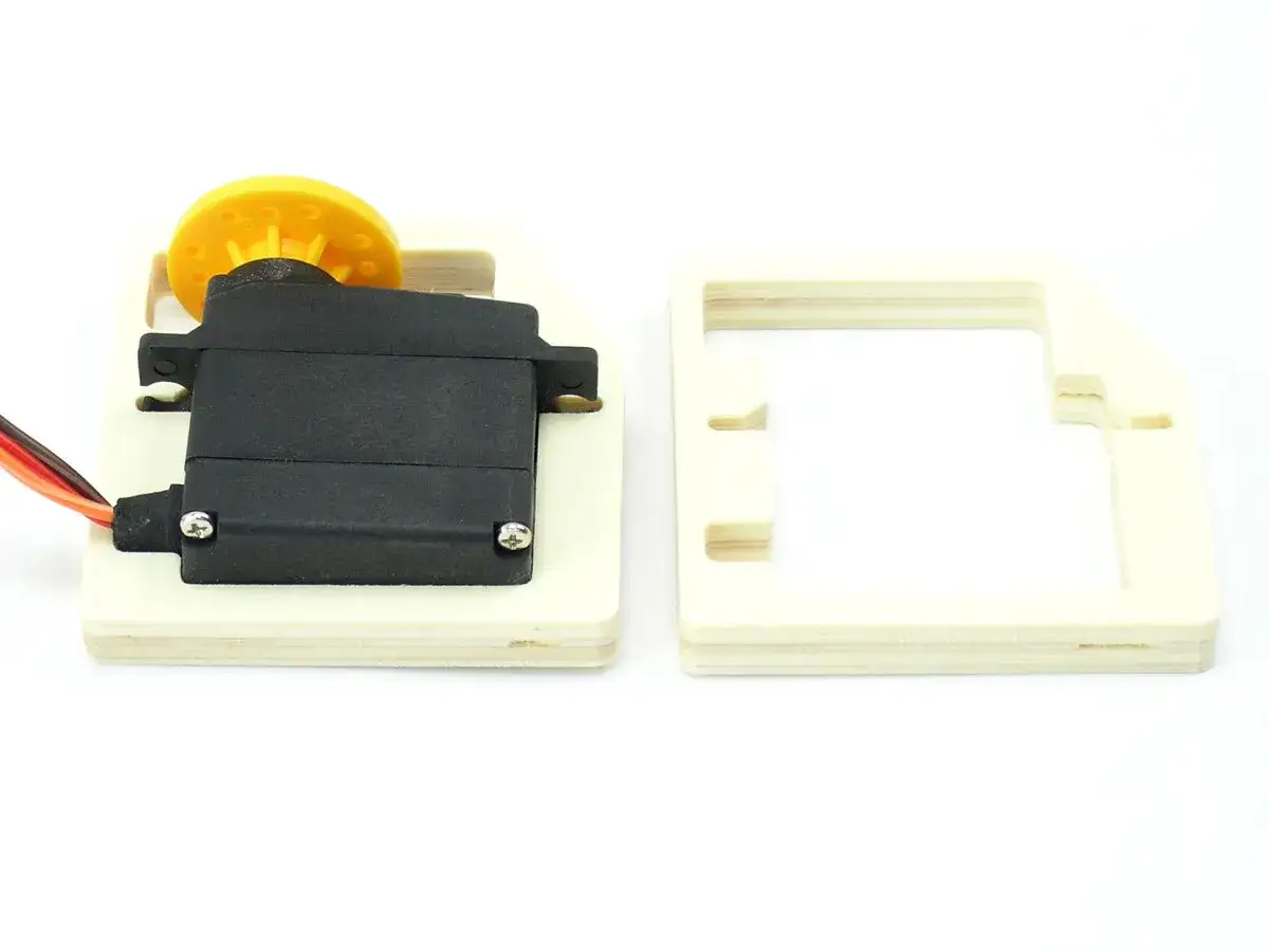 Ein Servomotor mit gelbem Rad auf Holz montiert, daneben ein Holzrahmen. Ein kleiner, pr&auml;ziser dymond-Mechanismus.