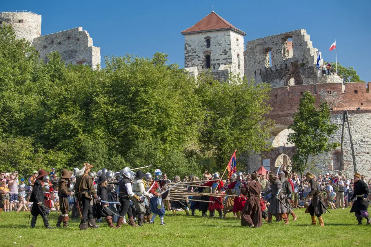 Zamek Tenczyn w Rudnie ożywa podczas inscenizacji historycznej. Grupa rekonstruktorów w strojach z epoki, z mieczami i tarczami, przygotowuje się do walki na tle ruin zamku.