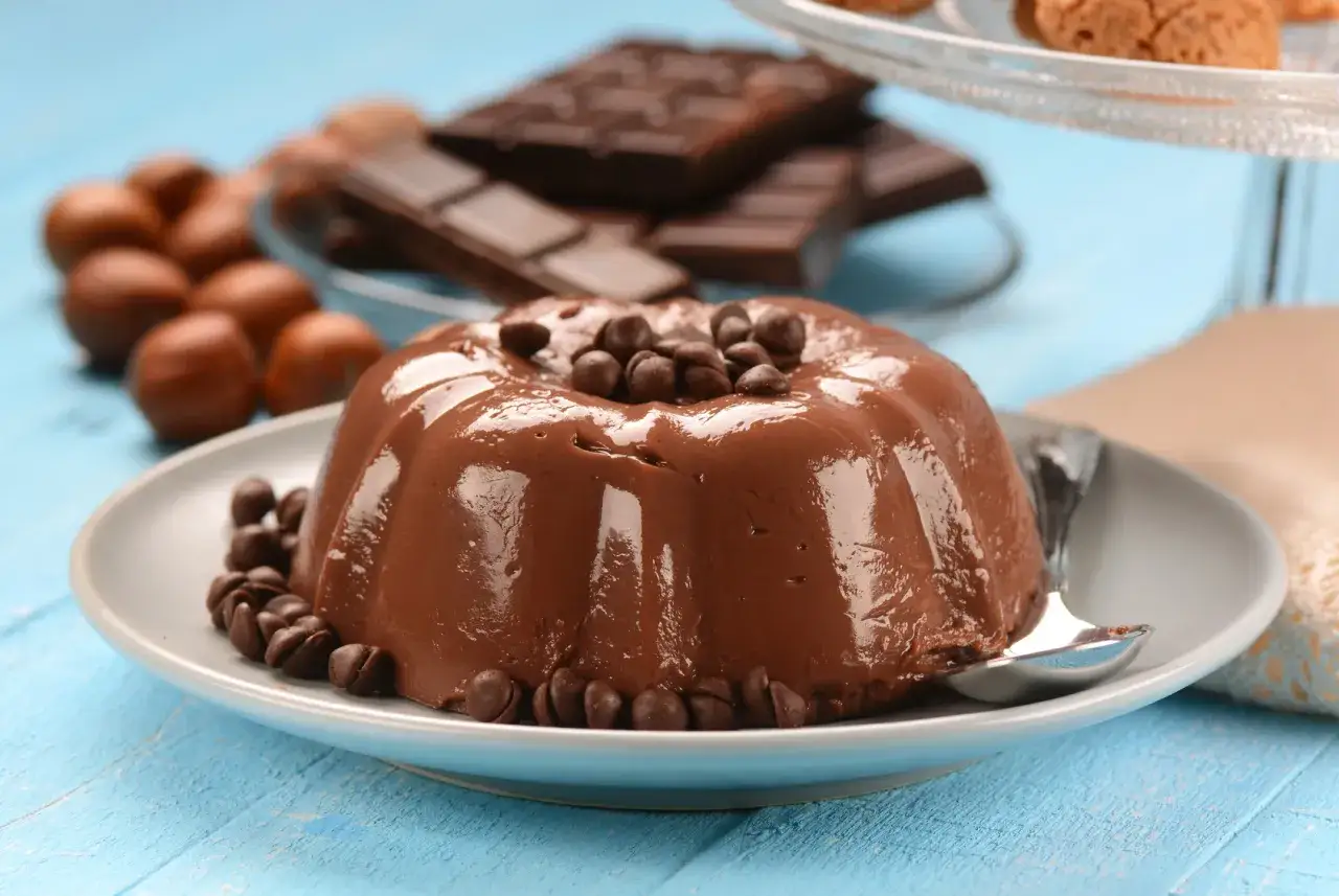Budino al cioccolato lucido, guarnito con gocce di cioccolato, su un piatto grigio. Sullo sfondo, tavolette di cioccolato e nocciole.