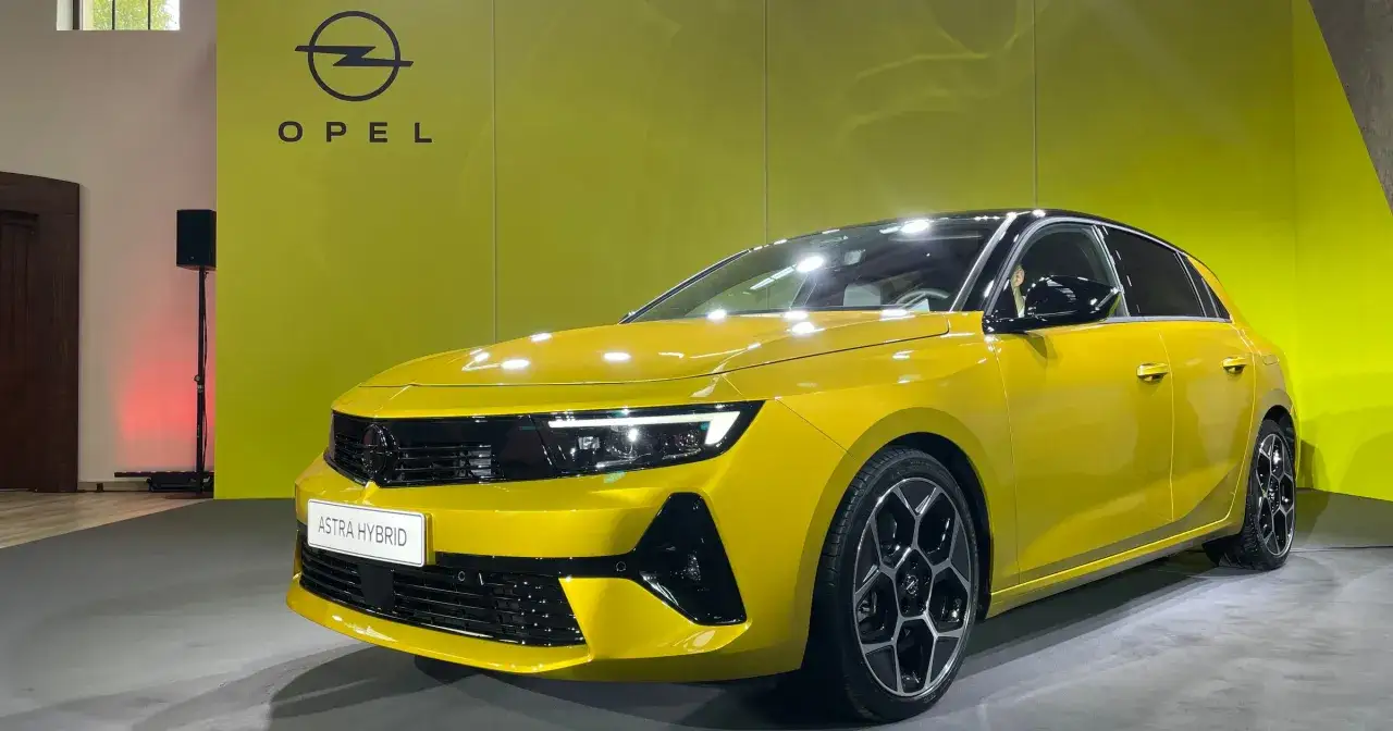 Nowy, żółty Opel Astra Hybrid prezentowany na tle logo marki. To jeden z najnowszych modeli Opla.