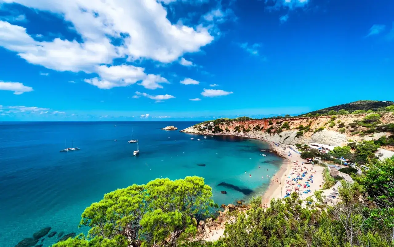 Piękna zatoka na Balearach, z turkusową wodą, plażą pełną ludzi i żaglówkami na morzu. Jakie wyspy Balearów oferują taki widok?