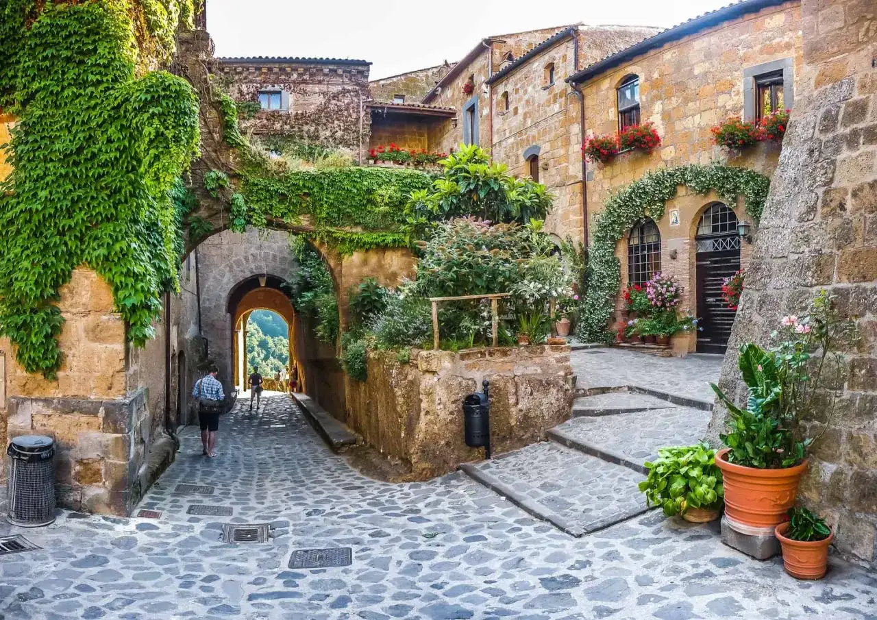 Un vicolo acciottolato con case in pietra ricoperte di edera e fiori. Un uomo cammina verso un arco, uno dei tanti scorci incantevoli tra le 10 cose da vedere nelle Marche.