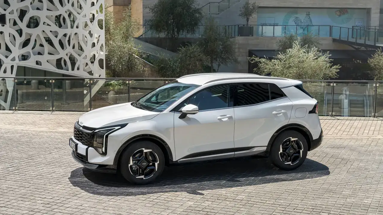 Biały SUV Kia Sportage, jeden z najnowszych modeli, zaparkowany na brukowanym placu przed nowoczesnym budynkiem z ażurową fasadą.
