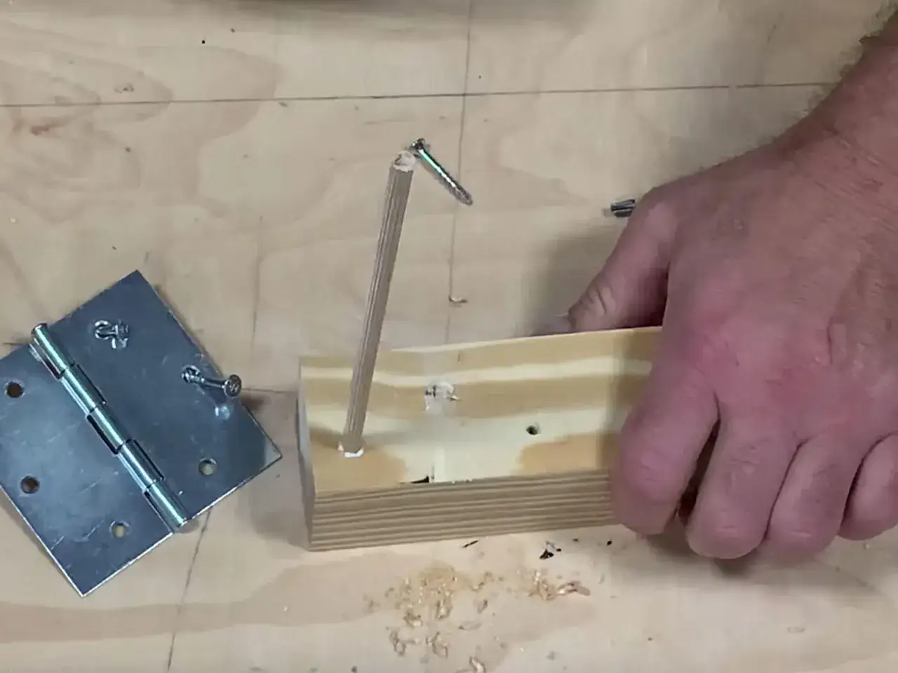 Un bricoleur teste un montage : une vis qui tourne dans le vide, tenue par un bout de bois, à côté d'une charnière.