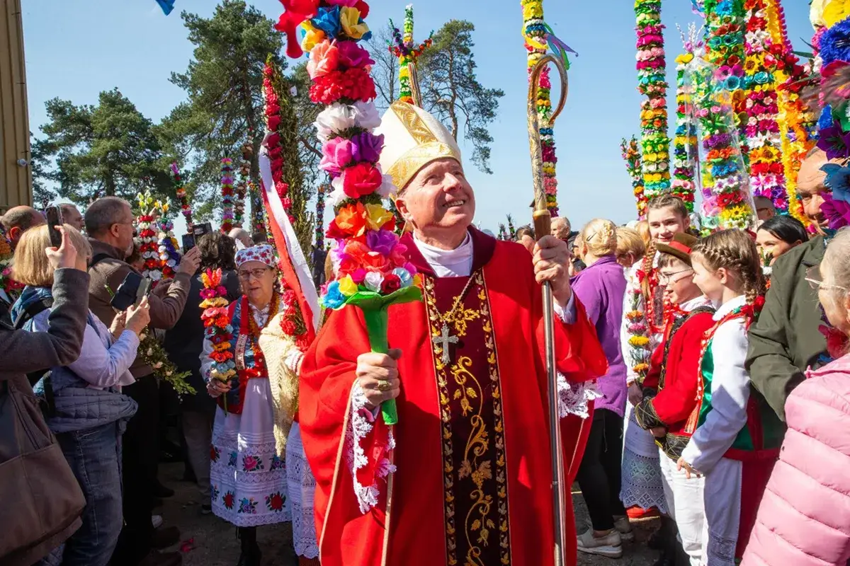 Biskup z barwną palmą w dłoni, otoczony ludźmi w tradycyjnych strojach, celebruje uroczystość na niedzielę palmową.