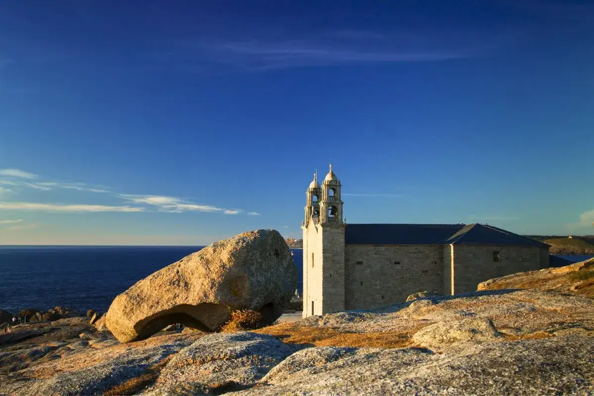 Iglesia de piedra junto al mar, con los penedos de pasarela e traba en primer plano.