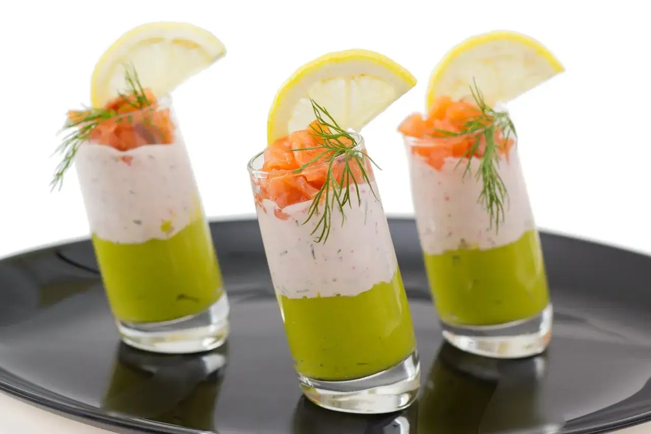 Trois verrines apéro colorées : avocat, crème et saumon fumé, garnies d'aneth et d'une tranche de citron.