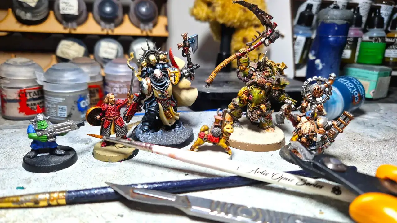 Miniature di Warhammer+ in preparazione: un orco, un guerriero, un demone e un nano. Pennelli e colori sullo sfondo.