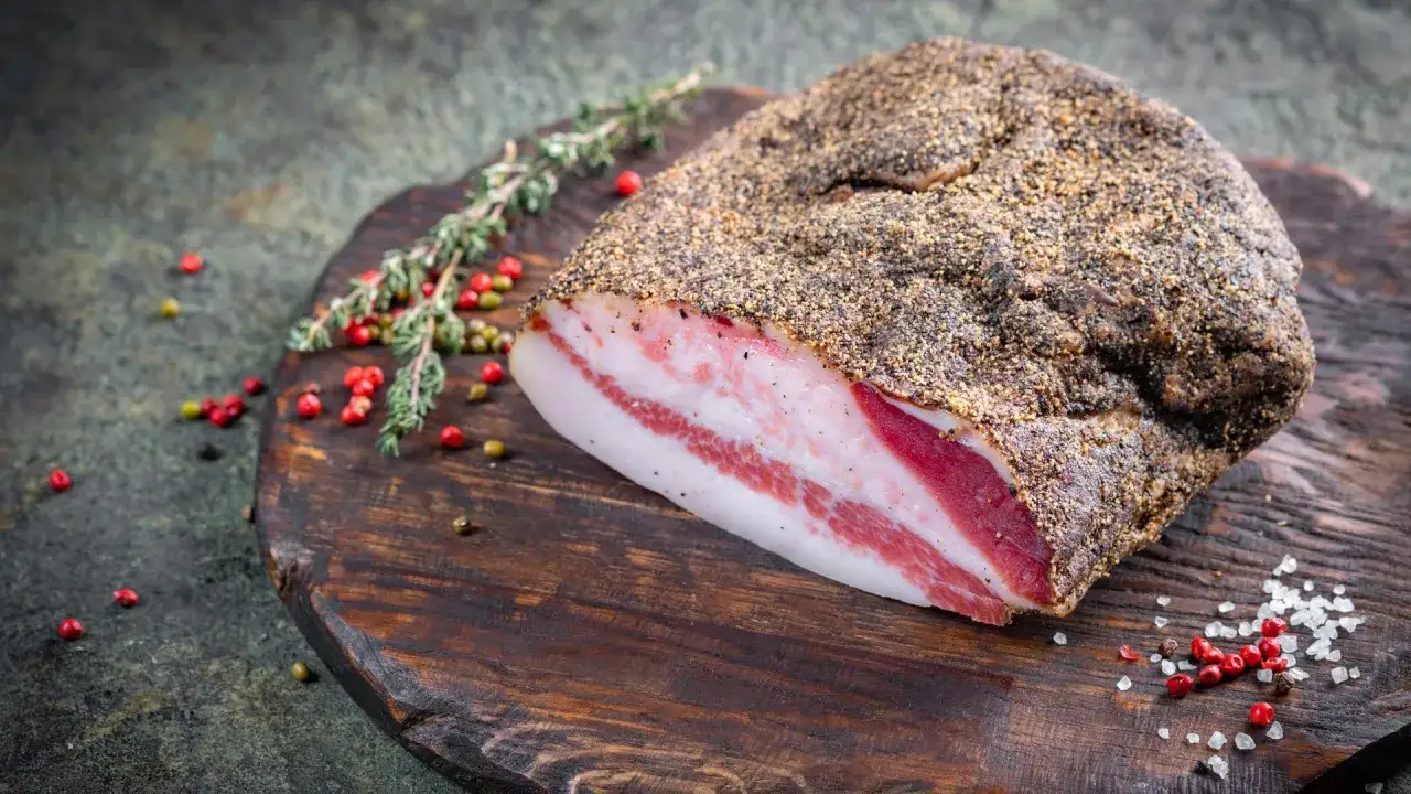 Guanciale co to? Wędzony kawałek wieprzowiny z widocznymi warstwami tłuszczu i mięsa, przyprawiony pieprzem, na drewnianej desce z ziołami i pieprzem.
