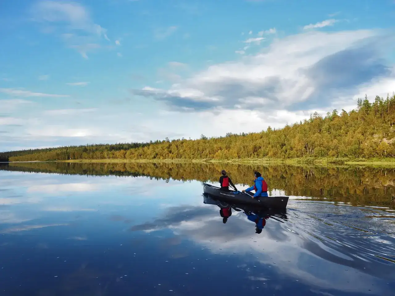Två personer paddlar kanot på en spegelblank sjö i Sverige. Skogen längs stranden speglas i vattnet.