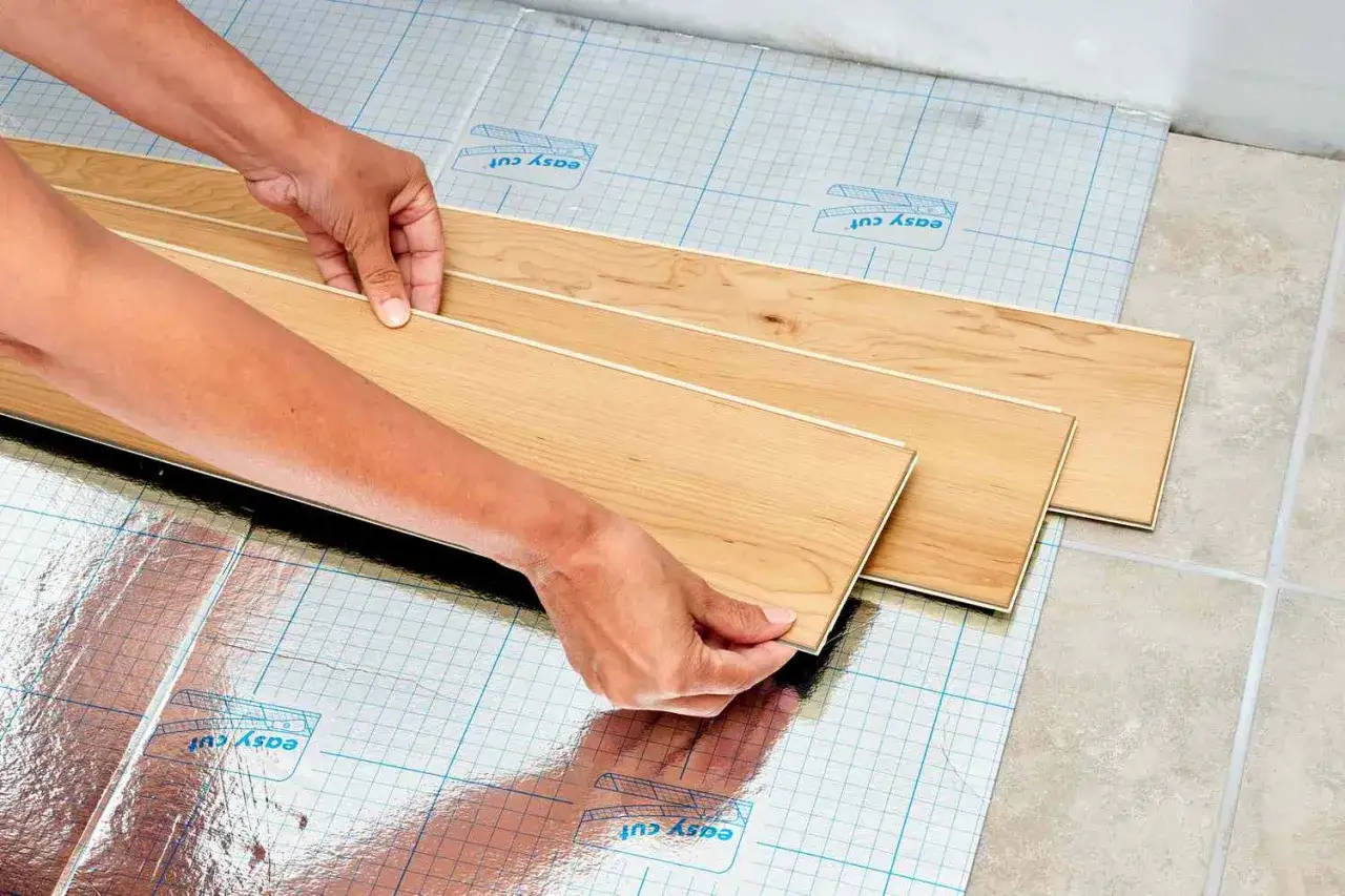 Hands lay down wood-look laminate flooring over a vinyl floor with a reflective underlayment.