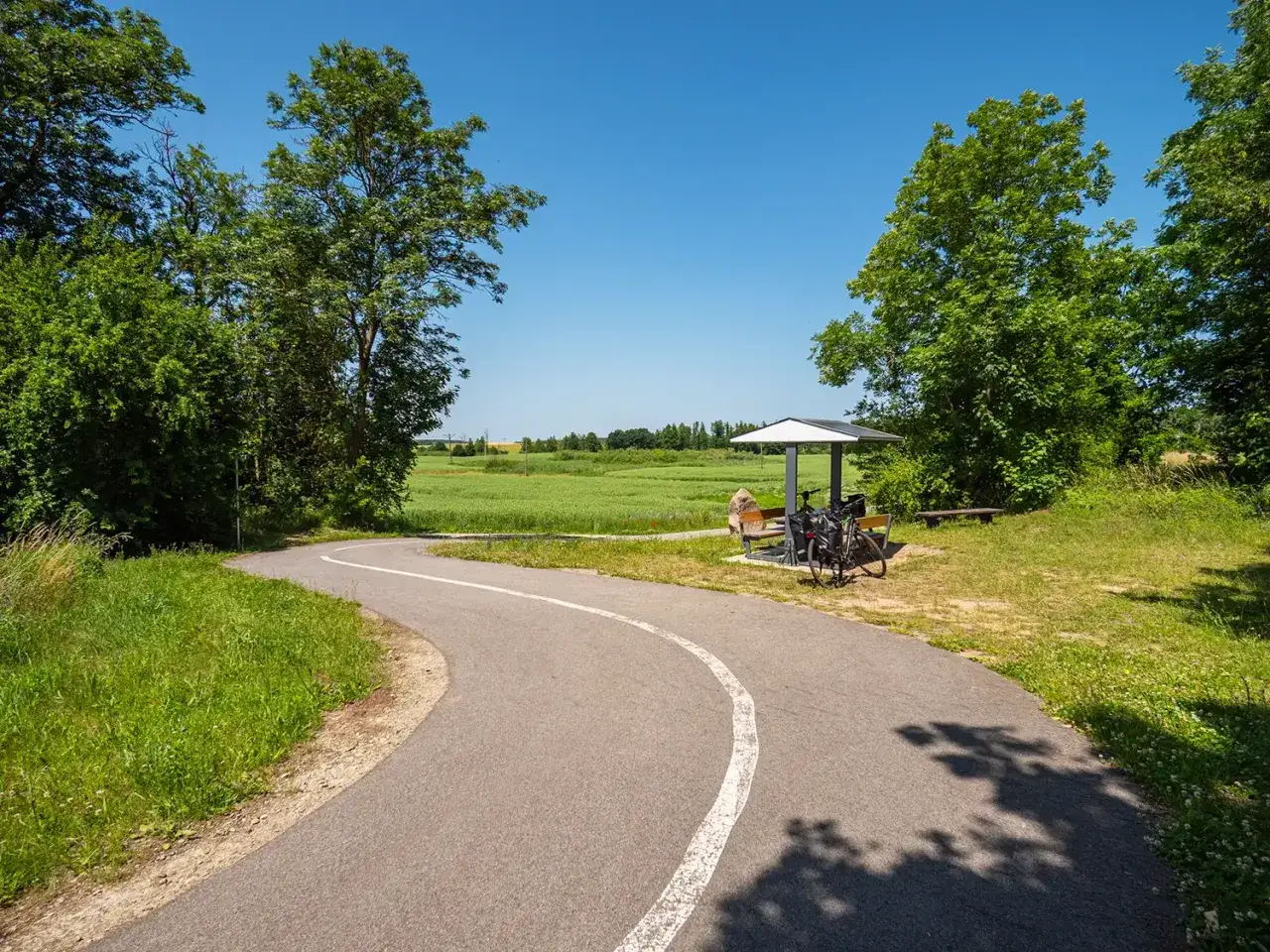Asfaltowa ścieżka rowerowa wije się przez zielone pola. Przy wiacie odpoczynek dla rowerzystów, idealne miejsce na przerwę podczas podróży trasą pojezierzy zachodnich.
