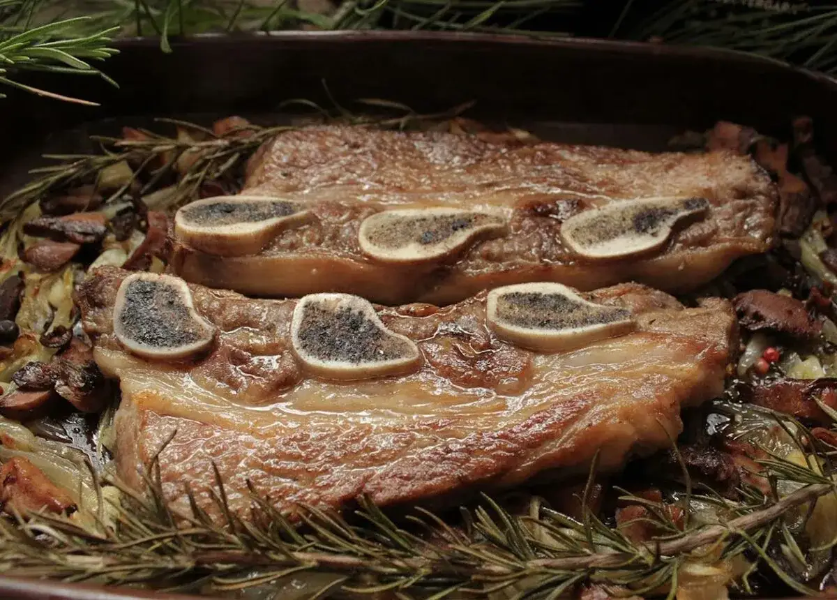 Dos piezas de redondo de ternera asado, con huesos expuestos, sobre una cama de cebolla, champi&ntilde;ones y romero.