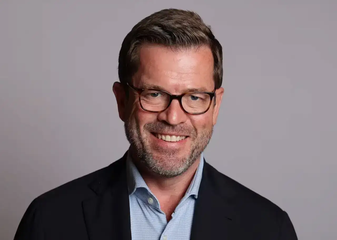 Portr&auml;t von Karl-Theodor zu Guttenberg mit Brille und Anzug, l&auml;chelnd vor grauem Hintergrund.