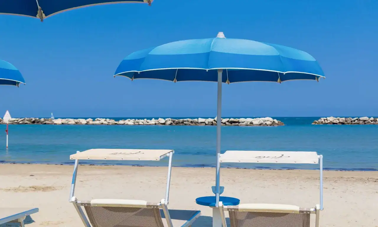 Ombrelloni blu e lettini sulla spiaggia, con il mare calmo e i frangiflutti in lontananza, tipici dei paesi vicino Rimini sul mare.