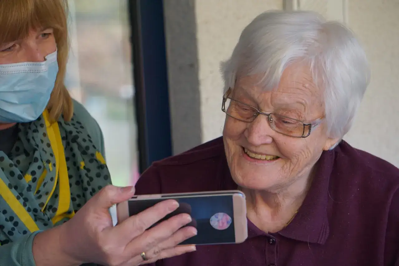 Una donna con mascherina mostra un telefono a un'anziana sorridente. L'accompagnamento anziani a chi spetta è un tema importante.
