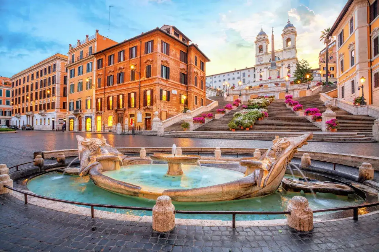 Fontana della Barcaccia e la scalinata di Trinit&agrave; dei Monti a Roma, con edifici storici e fiori colorati.