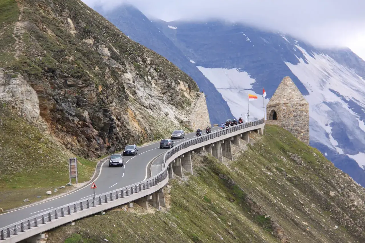 Malownicza trasa Grossglockner Hochalpenstrasse z krętymi drogami, samochodami i motocyklami, w tle majestatyczne g&oacute;ry z resztkami śniegu.