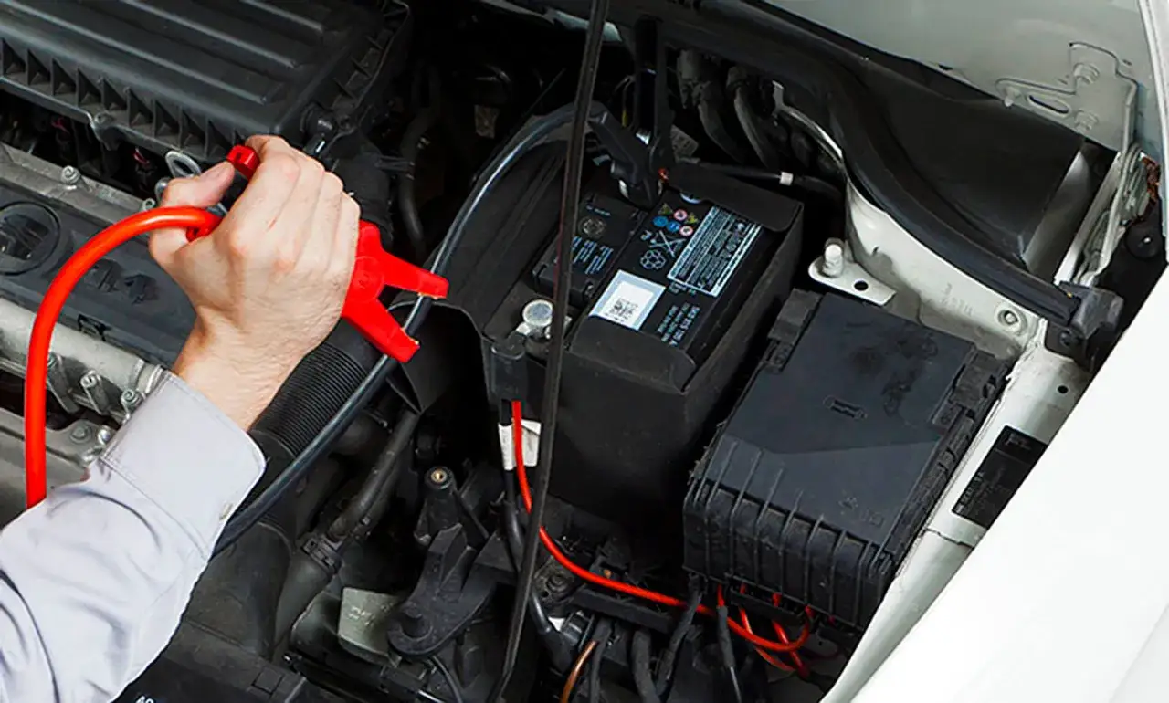 Mann schlie&szlig;t Starthilfekabel an die Autobatterie an. Rote Klemme am Pluspol, schwarze am Minuspol.