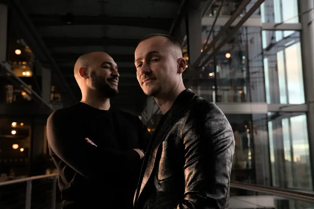 Zwei Männer, die die Musikindustrie repräsentieren, posieren in einem modernen Gebäude. Der Mann im Vordergrund trägt einen auffälligen Lederblazer.