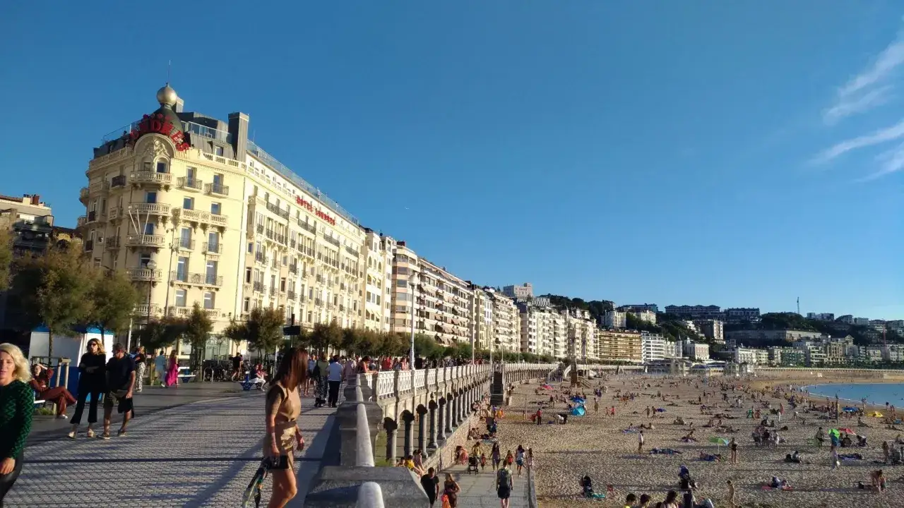 Gente paseando por el paseo mar&iacute;timo de la costa de San Sebasti&aacute;n, con la playa llena de ba&ntilde;istas y edificios hist&oacute;ricos al fondo.