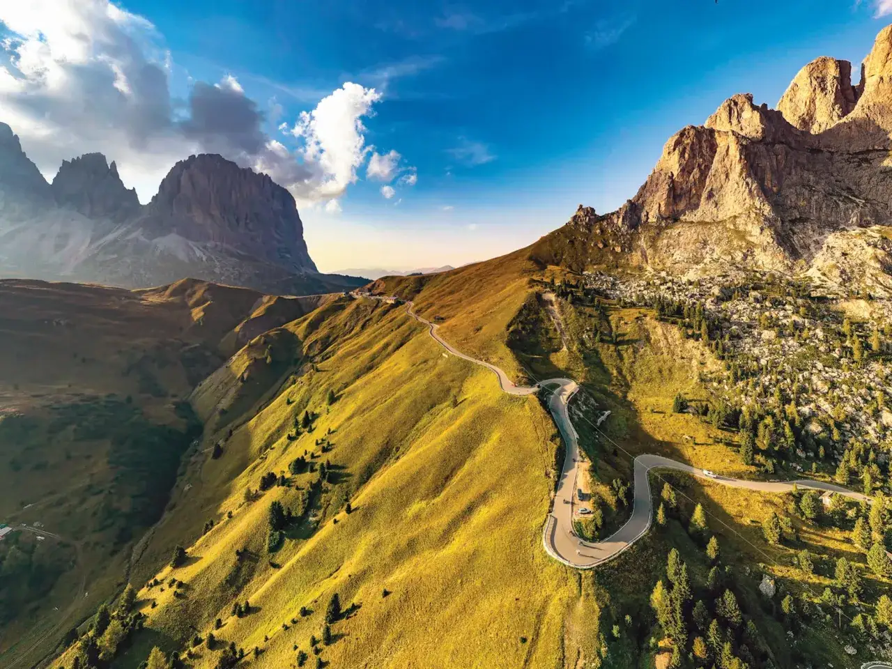 Słoneczna pogoda w Dolomitach. Kręta droga wijąca się pośród zielonych wzgórz, z majestatycznymi szczytami w tle.