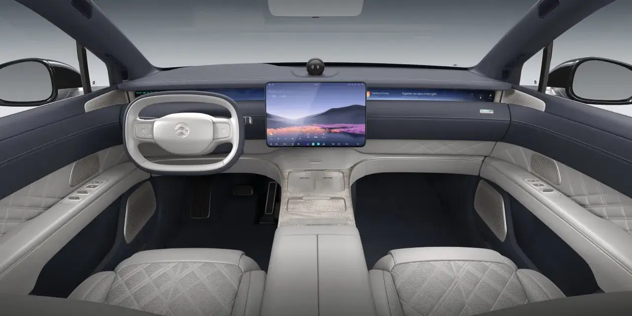 Interno lussuoso del NIO ET9 con sedili in pelle trapuntata, volante moderno e ampio display centrale che mostra paesaggi e musica.