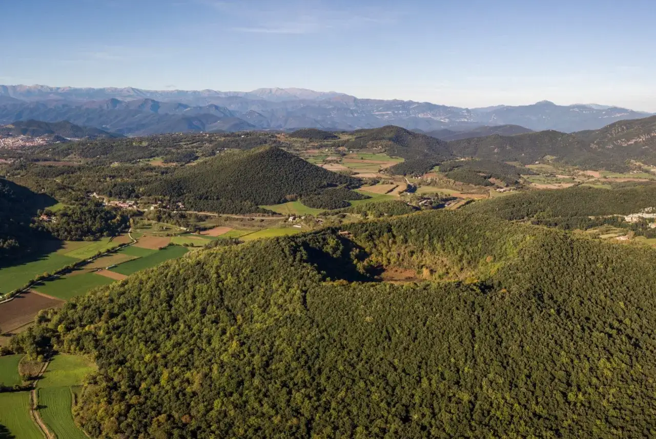 Paisaje aéreo de la ruta volcanes Garrotxa, con colinas verdes cubiertas de árboles y un cráter visible.