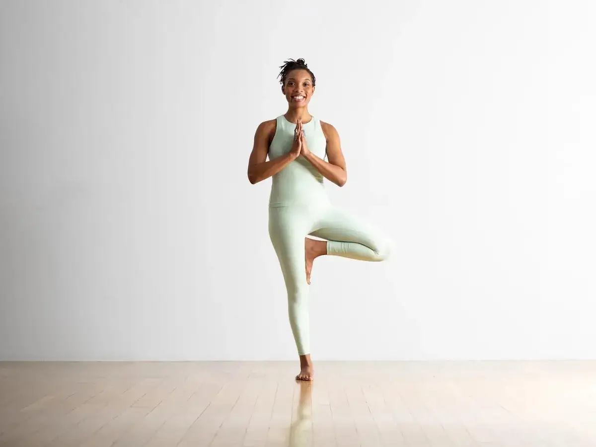 Mujer sonriente en postura del arbol yoga, con el pie derecho apoyado en el muslo izquierdo.