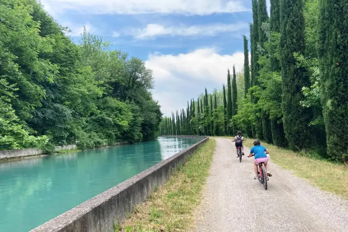 Due bambini pedalano lungo la ciclovia del Mincio, costeggiando un canale dalle acque turchesi e un viale di cipressi.