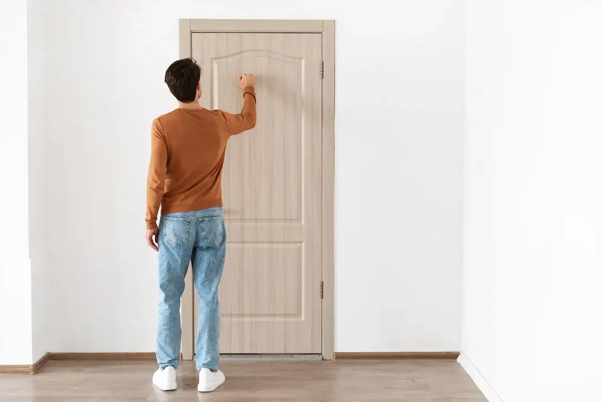 Uomo con maglione arancione e jeans bussa a una porta interna in legno chiaro, un esempio di montaggio porte interne.