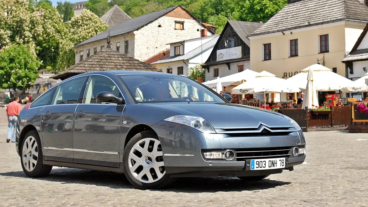 Szary Citroën C6 na brukowanym rynku, z widokiem na urokliwe kamienne budynki i ogródek kawiarniany.