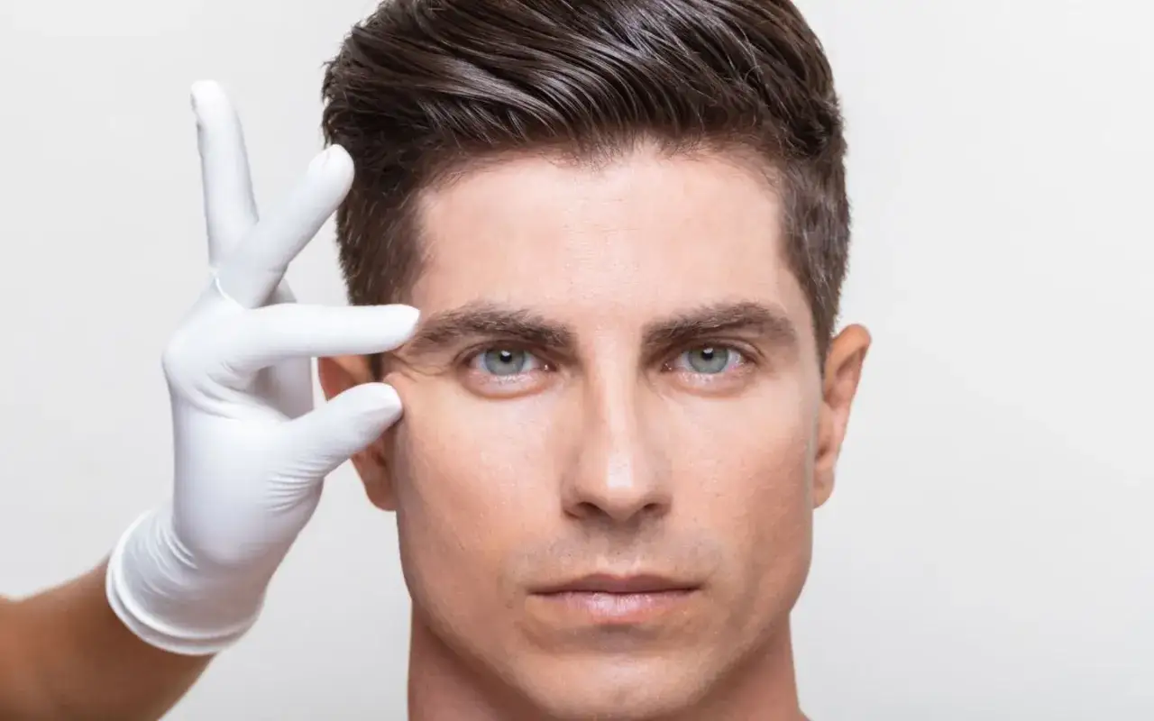 Main d'un professionnel en gant blanc tirant la peau du visage d'un homme pour examiner les rides frontales.