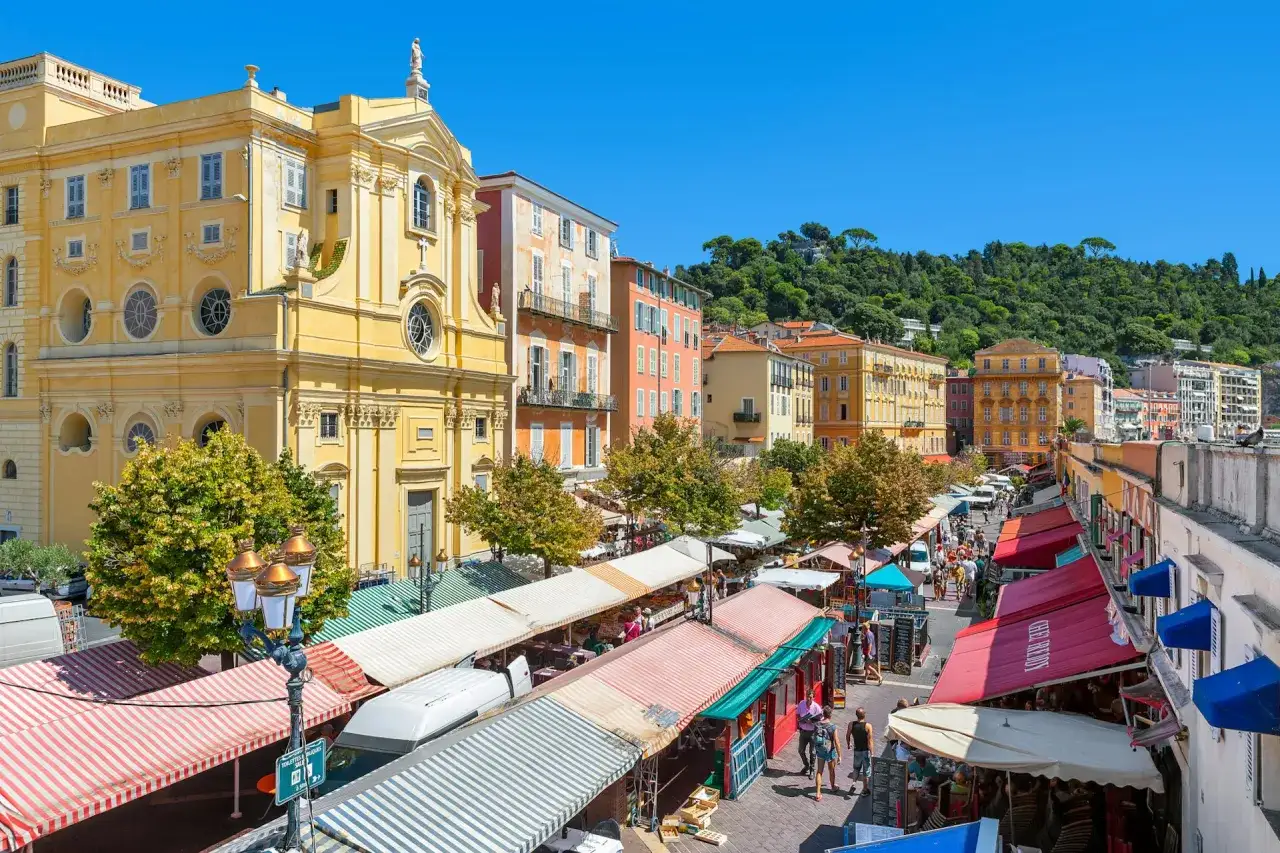 Tętniący życiem targ na Cours Saleya w Nicei, z kolorowymi straganami, zabytkową architekturą i zielonym wzgórzem w tle.