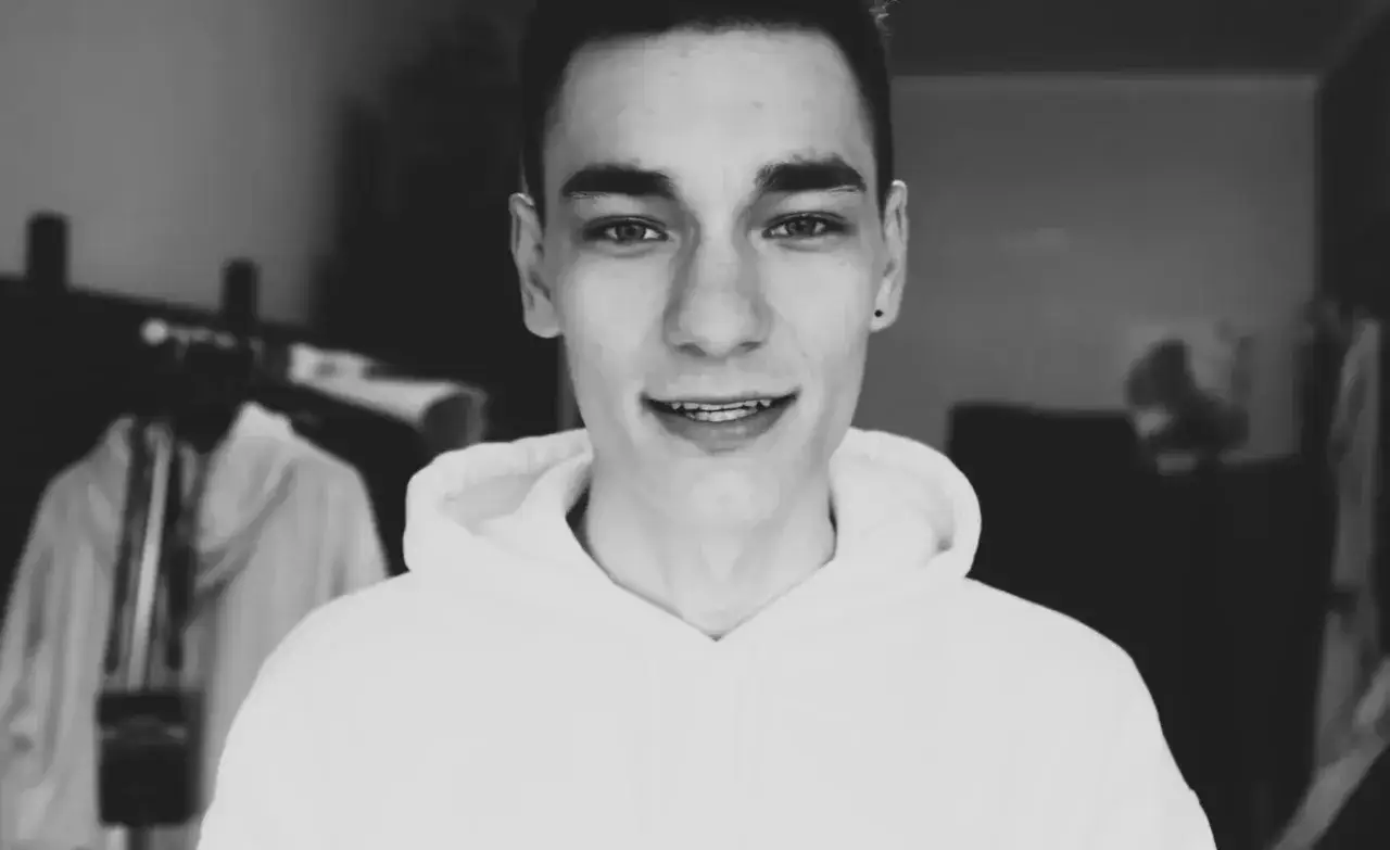 Uśmiechnięty chłopak, chati youtuber, w białej bluzie z kapturem, w tle widać inne osoby.