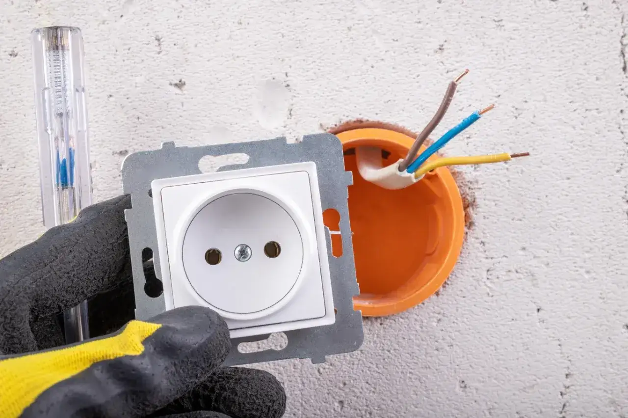 Ręka w rękawicy trzyma białe gniazdko, obok widać instalację elektryczną i próchnik. Dowiedz się, jak zrobić uziemienie w gniazdku.