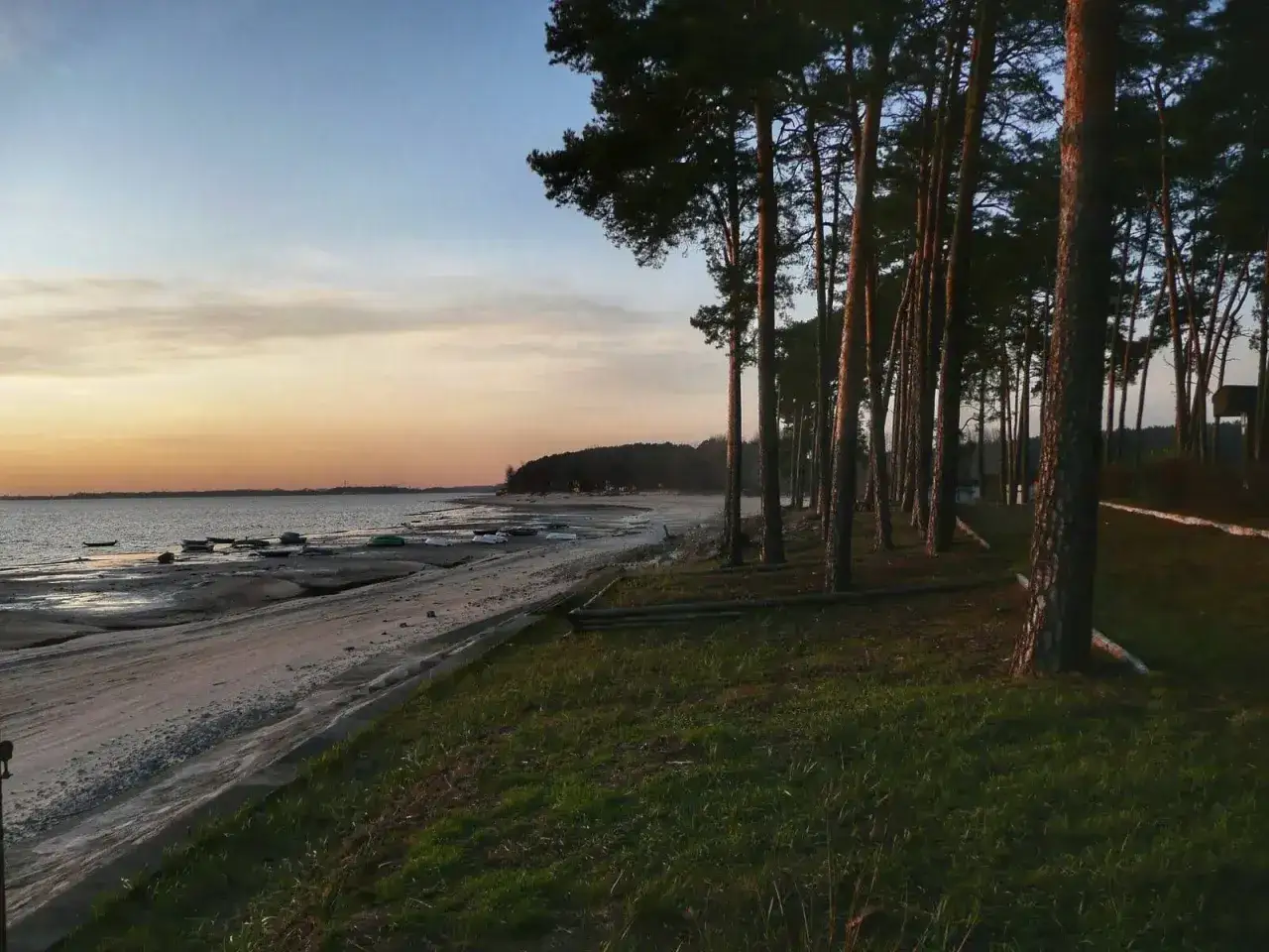 Zachód słońca nad otmuchowskim jeziorem. Rząd sosen wzdłuż piaszczystej plaży z łodziami.