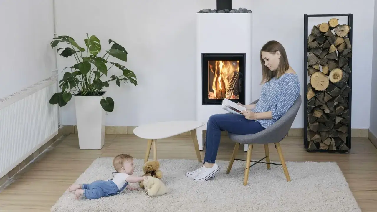Kobieta czyta książkę przy nowoczesnym, wolnostojącym kominku. Obok bawi się dziecko.