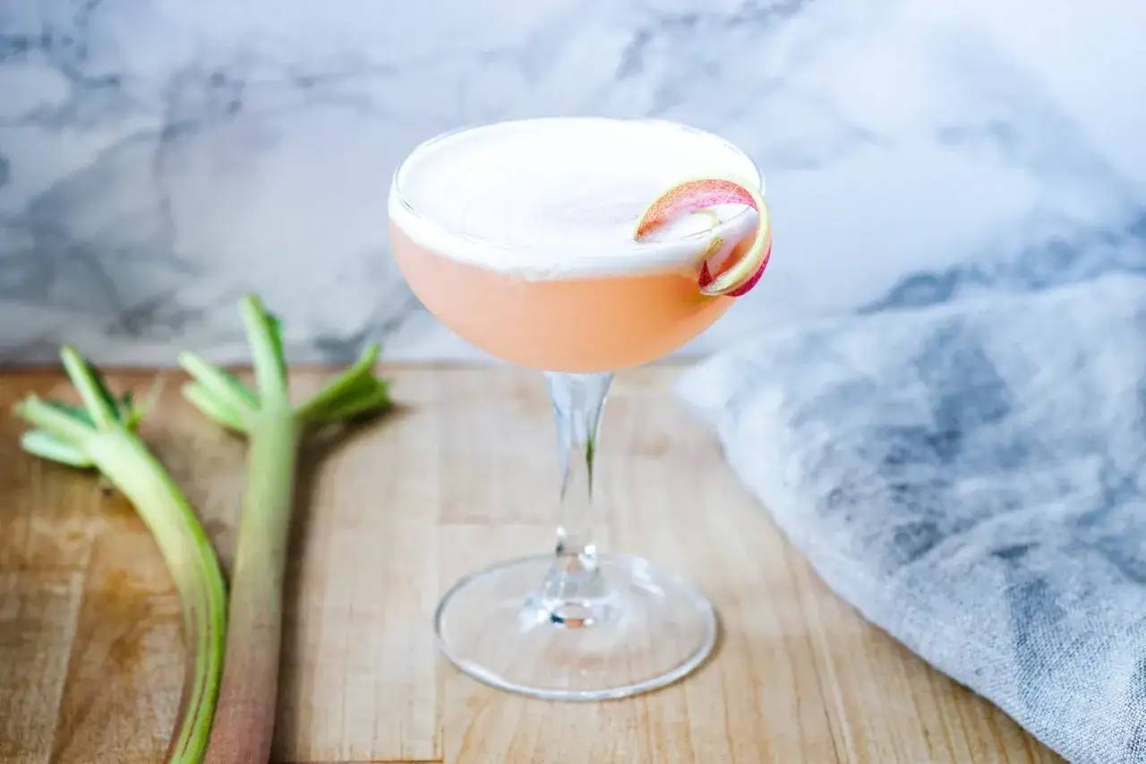 En rosa rabarber sour-drink med skum och en rabarberdekoration. Två rabarberstjälkar ligger bredvid.