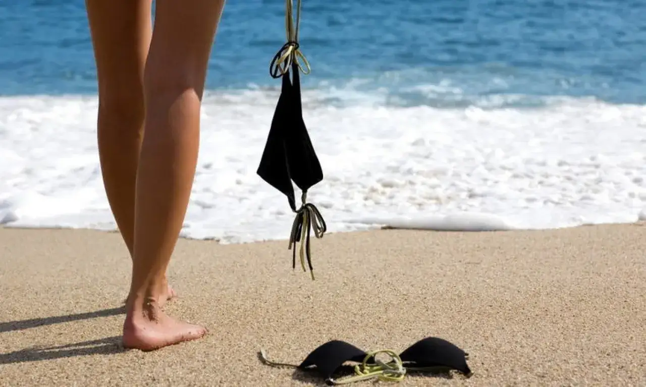 Nogi na piasku, obok czarny kostium kąpielowy. W tle morze i fale. Idealne miejsce na plażę nudystów w Polsce.