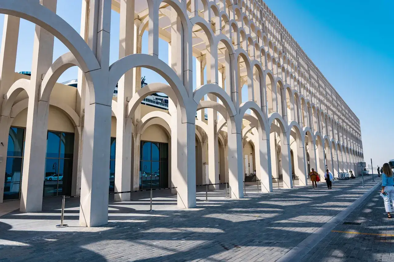 Ein langer Arkadengang mit vielen Bögen am doha port. Menschen spazieren auf dem gepflasterten Weg entlang der modernen Architektur.