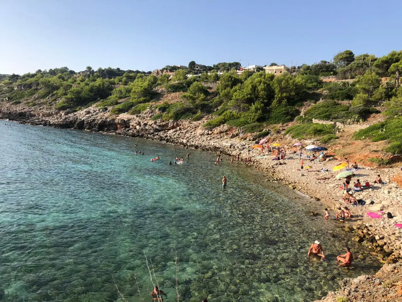 Spiaggia di San Gregorio Leuca con gente che nuota e prende il sole tra scogli e vegetazione mediterranea.