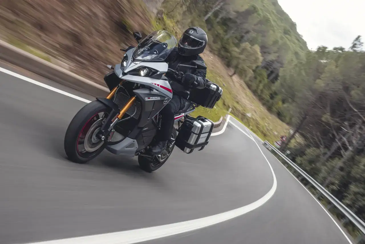 Motociclista con casco negro en una moto deportiva gris y plateada, con alforjas, en una carretera sinuosa rodeada de vegetación. Una **energíca experia** en movimiento.