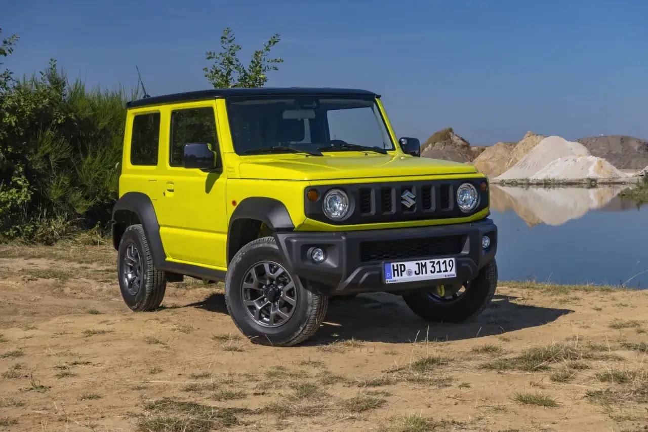 Żółty Suzuki Jimny, uznawany za najlepszy samochód terenowy, stoi na piaszczystym podłożu przy wodzie.