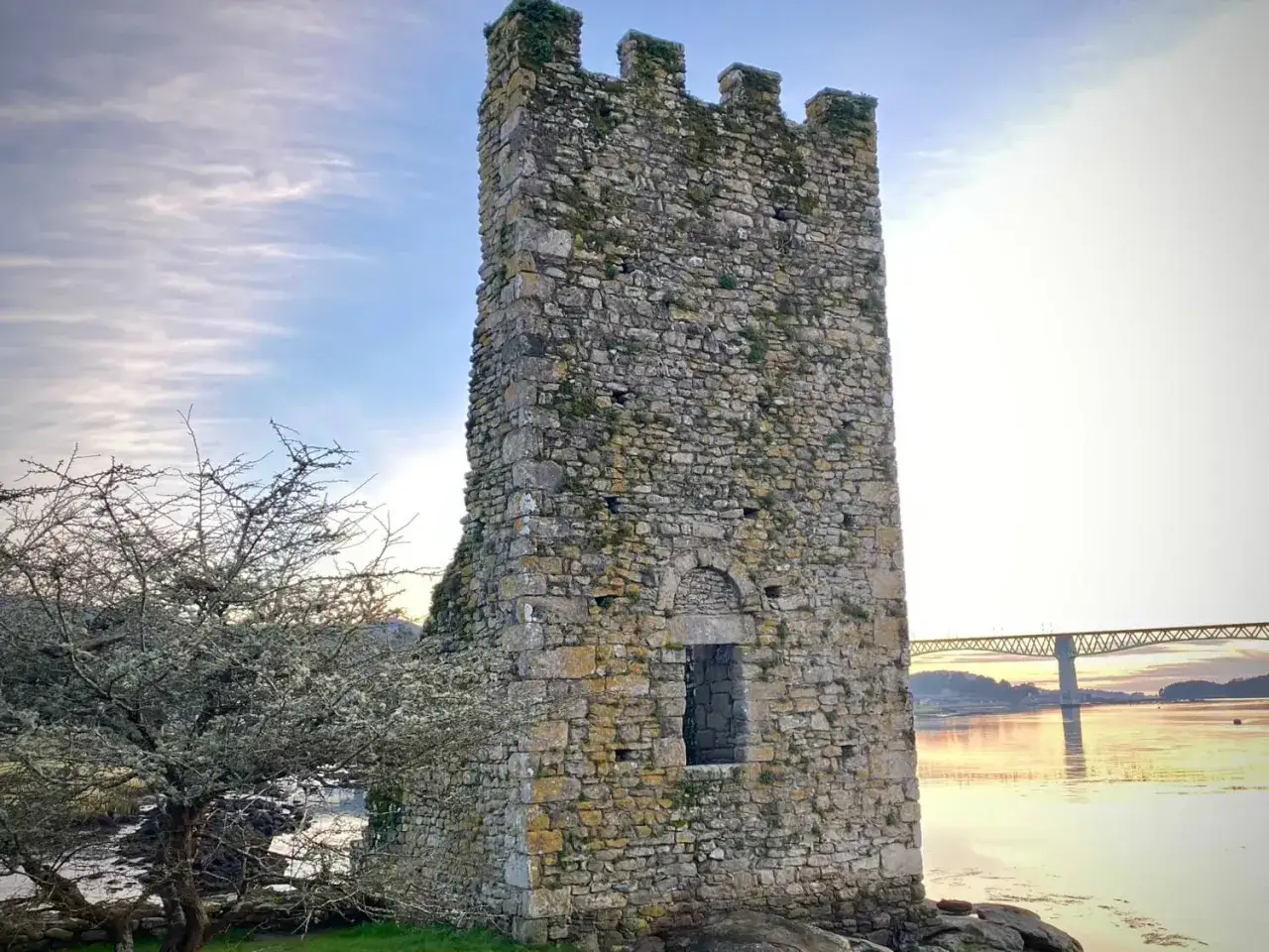 Torre medieval en Catoira, Galicia, con un puente al fondo reflejado en el agua.